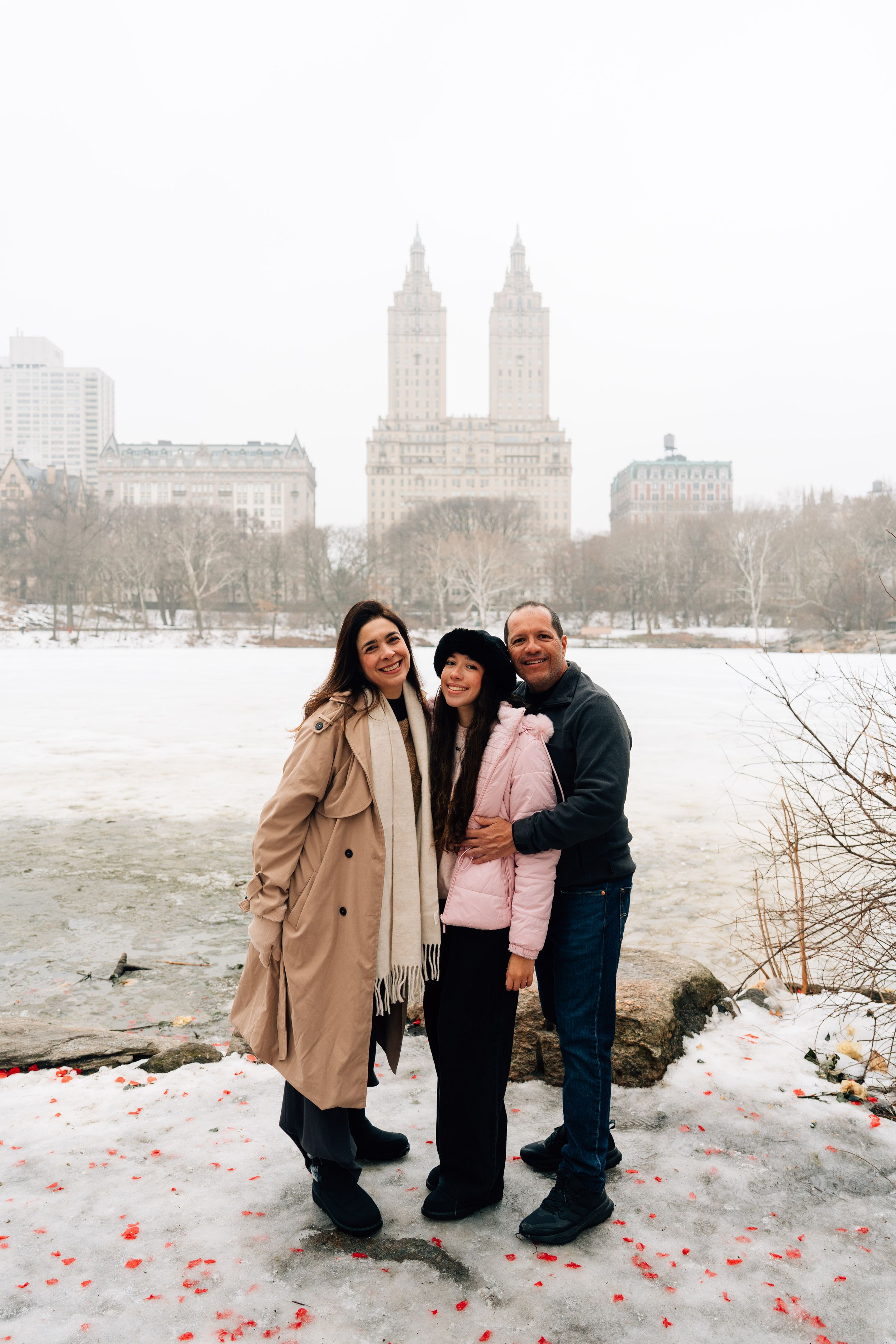 Three people pose smiling in snowy Central Park with a frozen lake and twin-towered building in the background.