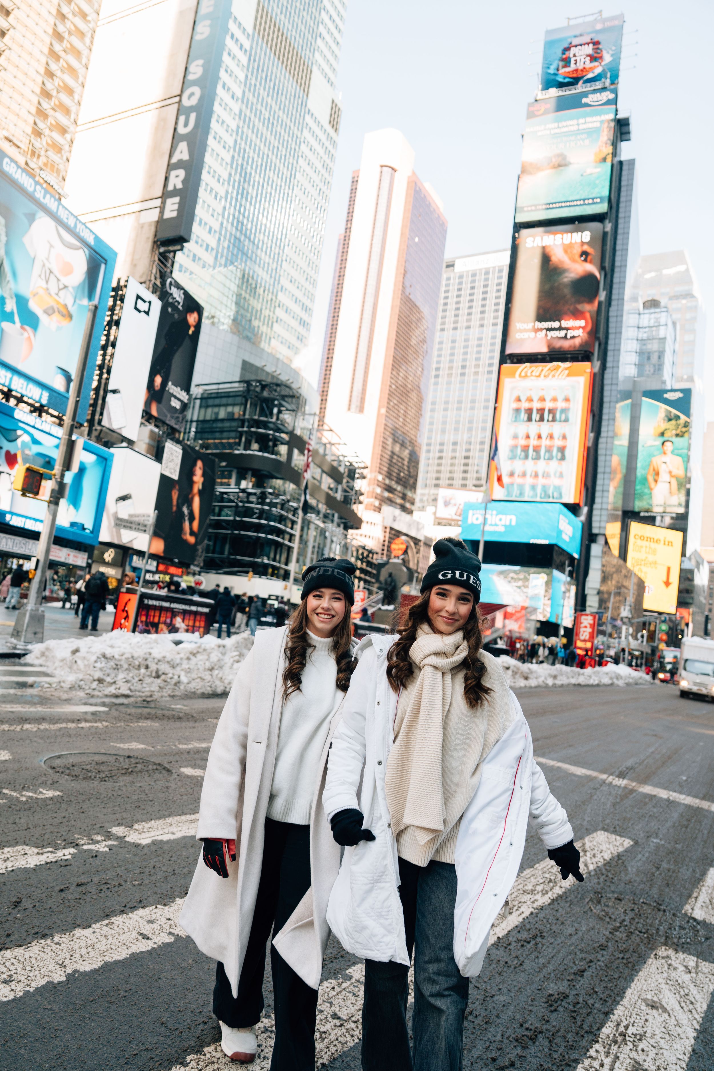 Two women in white winter coats and black beanies smiling in Times Square, New York City on a snowy day.