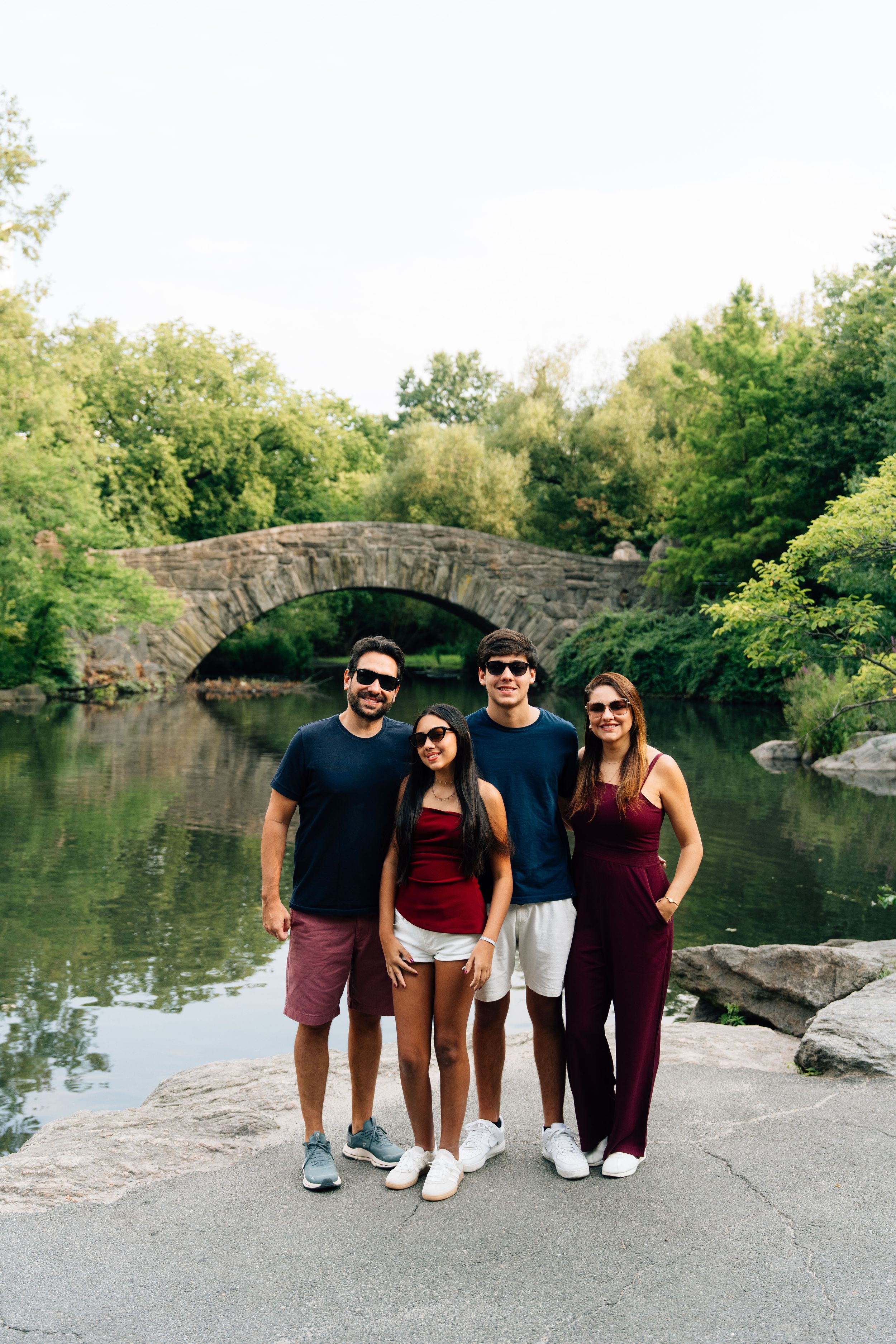 Four friends pose smiling by a stone arch bridge over a calm pond in Central Park, NYC, surrounded by lush green trees.