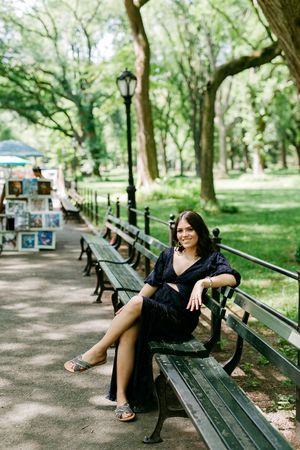 Person relaxing on park bench along tree-lined Central Park pathway with art vendor in background.