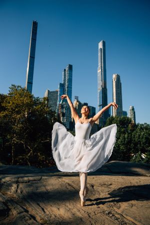 Ballerina in white dress performs graceful pose against NYC skyline with luxury towers.