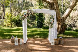 Elegant outdoor wedding arch adorned with white draping and pastel floral arrangements on a brick patio surrounded by lush greenery.