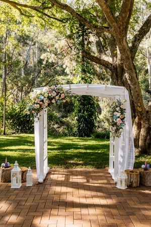 Elegant outdoor wedding arch adorned with pink and white flowers, white draping, surrounded by lush greenery on a brick pathway.