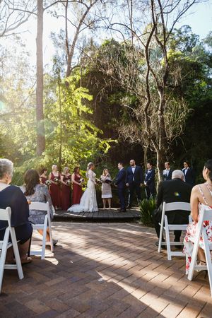 Outdoor wedding ceremony with bridesmaids in burgundy dresses, couple at altar surrounded by lush greenery and white chairs.