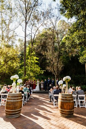Outdoor wedding ceremony on brick patio with wine barrel aisle markers, white floral arrangements, and lush green trees backdrop.