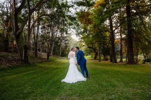 Bride and groom share a romantic kiss surrounded by lush green trees in an outdoor garden wedding setting.
