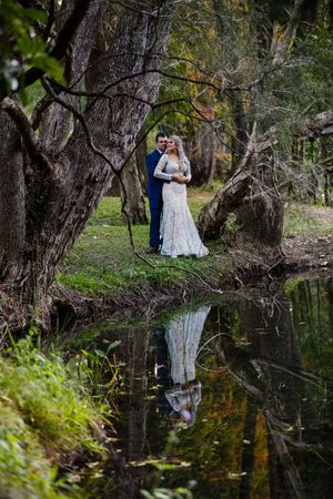 Bride and groom embracing by a reflective pond surrounded by lush trees in a romantic outdoor wedding portrait.