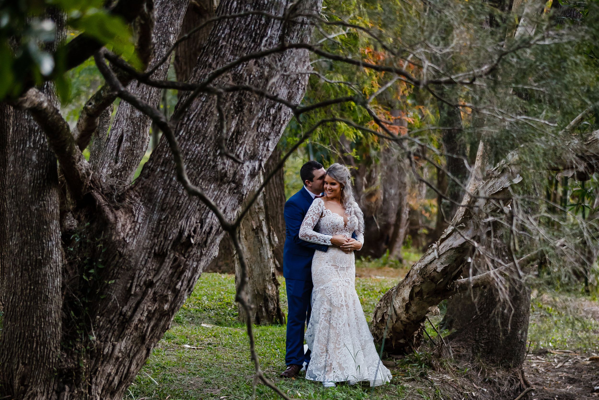 A bride and groom share a romantic embrace among ancient twisted trees in a lush forest wedding photo.