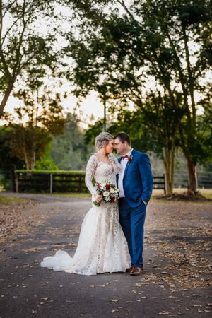 Bride in lace gown and groom in blue suit share a romantic moment on a tree-lined path at golden hour.