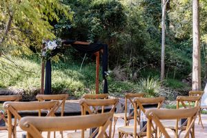 Outdoor wedding ceremony setup with wooden chairs, dark floral arch, and lush green forest backdrop.