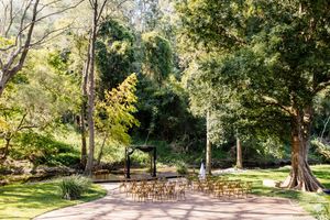 Outdoor wedding ceremony setup with wooden chairs and floral arch surrounded by lush green trees in a garden setting.