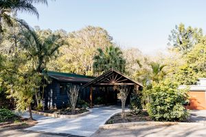 Dark blue cabin-style home with gabled carport, lush tropical gardens, and tall eucalyptus trees in a serene Australian setting.