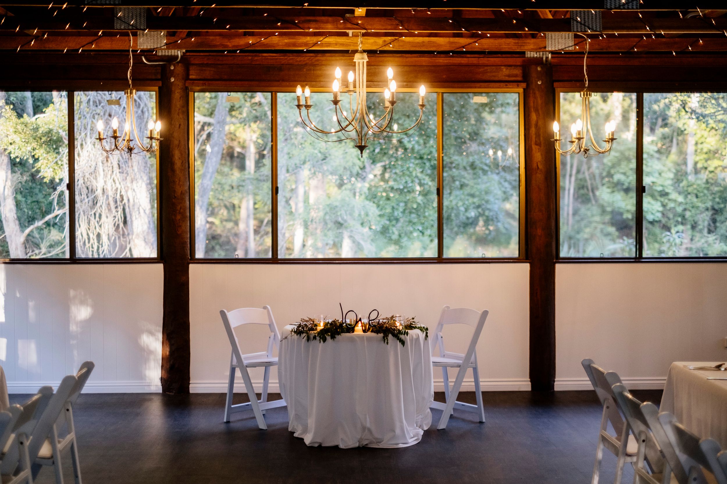 Elegant wedding sweetheart table with white linen, floral centerpiece, and chandeliers by large windows.