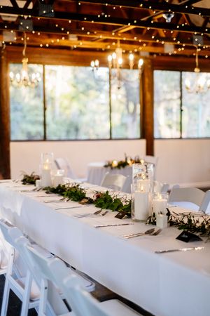Elegant wedding reception table with white linens, greenery garland, candles, and string lights in a rustic venue.