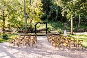 Outdoor wedding ceremony setup with wooden cross-back chairs, floral arch, and lush green trees in a garden setting.