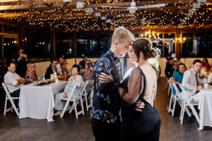 Couple shares first dance at wedding reception with string lights and guests seated at white tables in background.