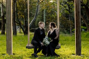Couple in black attire sitting on a wooden swing in a lush green garden, bride holding white bouquet.