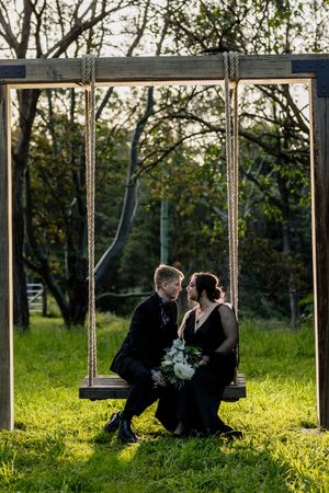 Couple in black wedding attire sharing a romantic moment on a wooden swing surrounded by lush green trees.