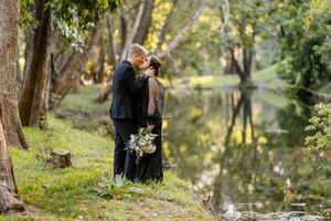 Couple sharing a romantic kiss in black wedding attire by a serene forest river, bride holding a white bouquet.