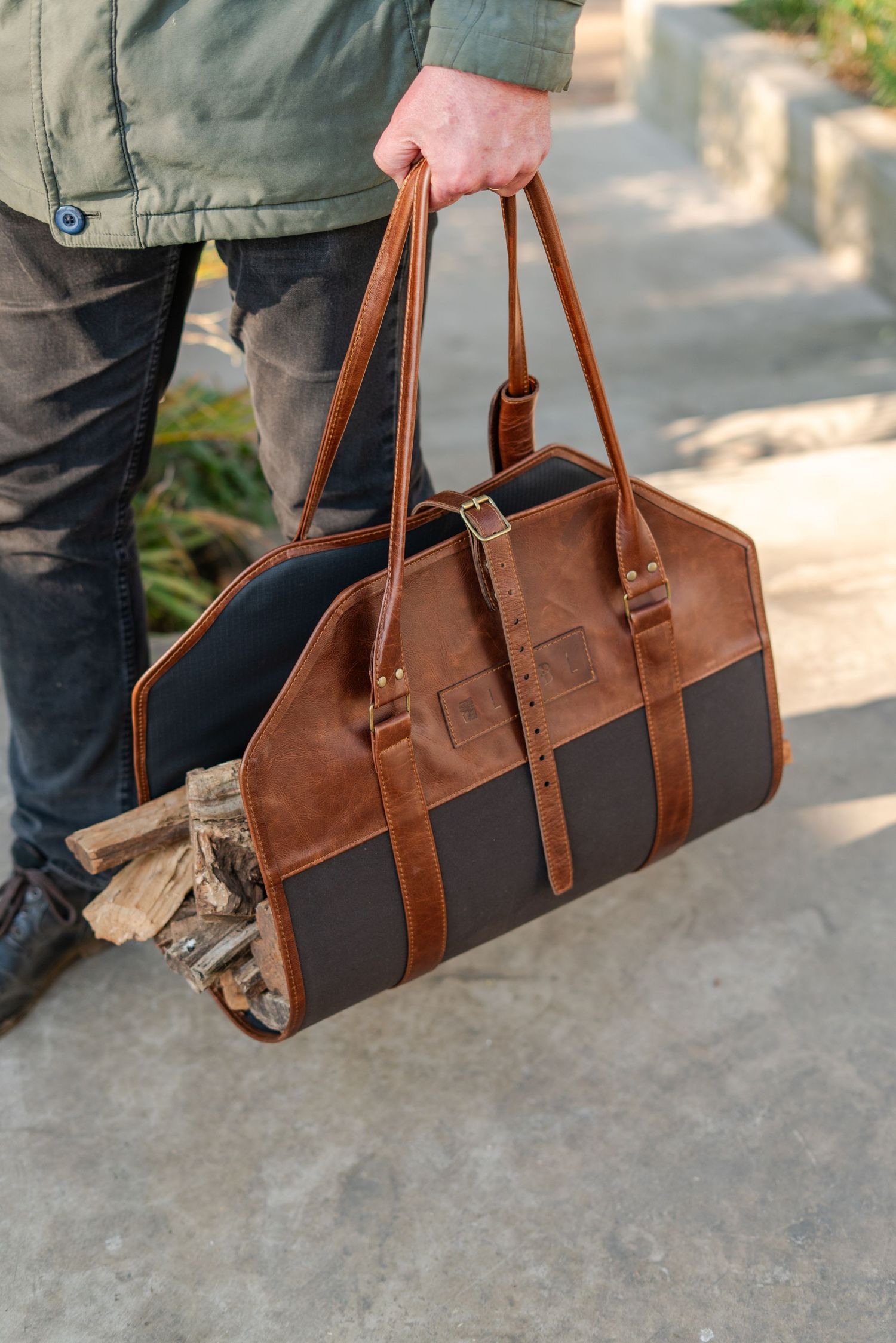 Man carrying a brown leather LYBL log carrier bag with firewood, featuring dual handles and dark canvas panels.