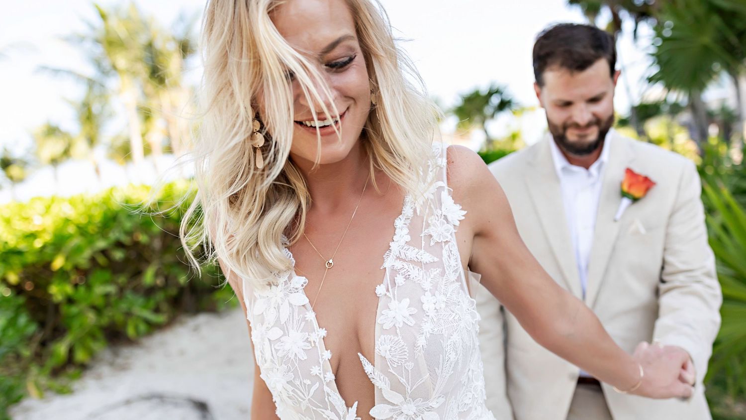 Bride in lace wedding gown smiles as groom in tan suit with orange boutonniere holds her hand on tropical beach.