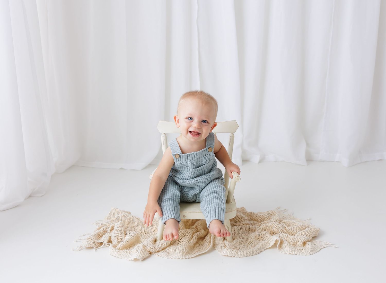 Smiling baby in blue knit romper sitting on white chair with beige blanket in bright white studio.