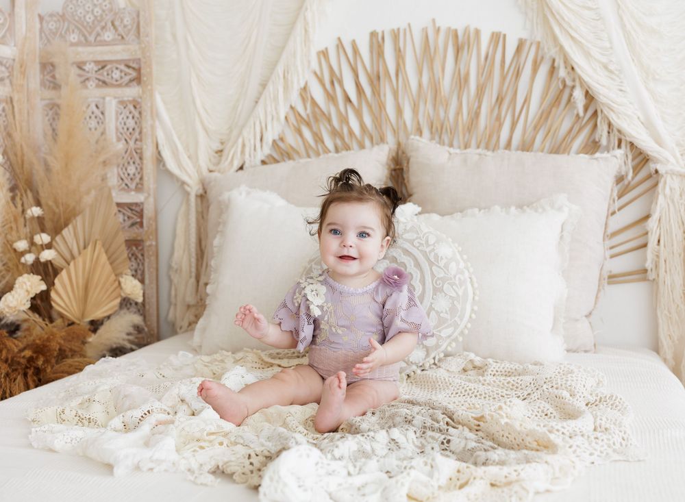 Baby girl in purple romper sits on boho-styled bed with white pillows and macrame blanket in dreamy nursery setting.