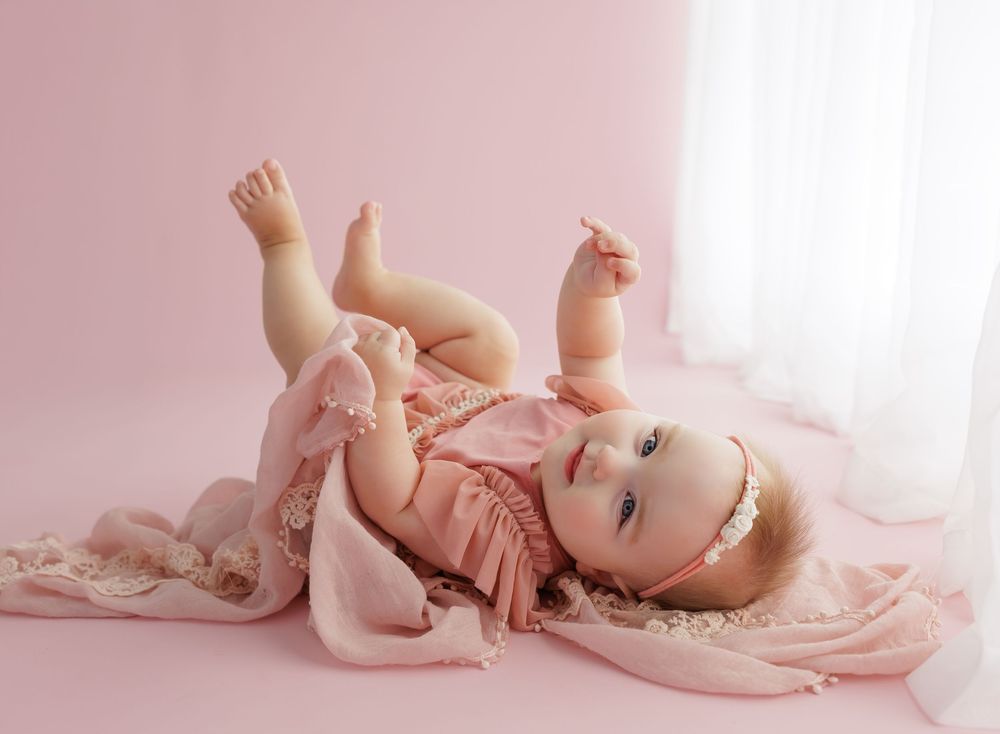 A baby girl in a pink romper lies on a pink backdrop, playfully lifting her legs and looking at the camera.