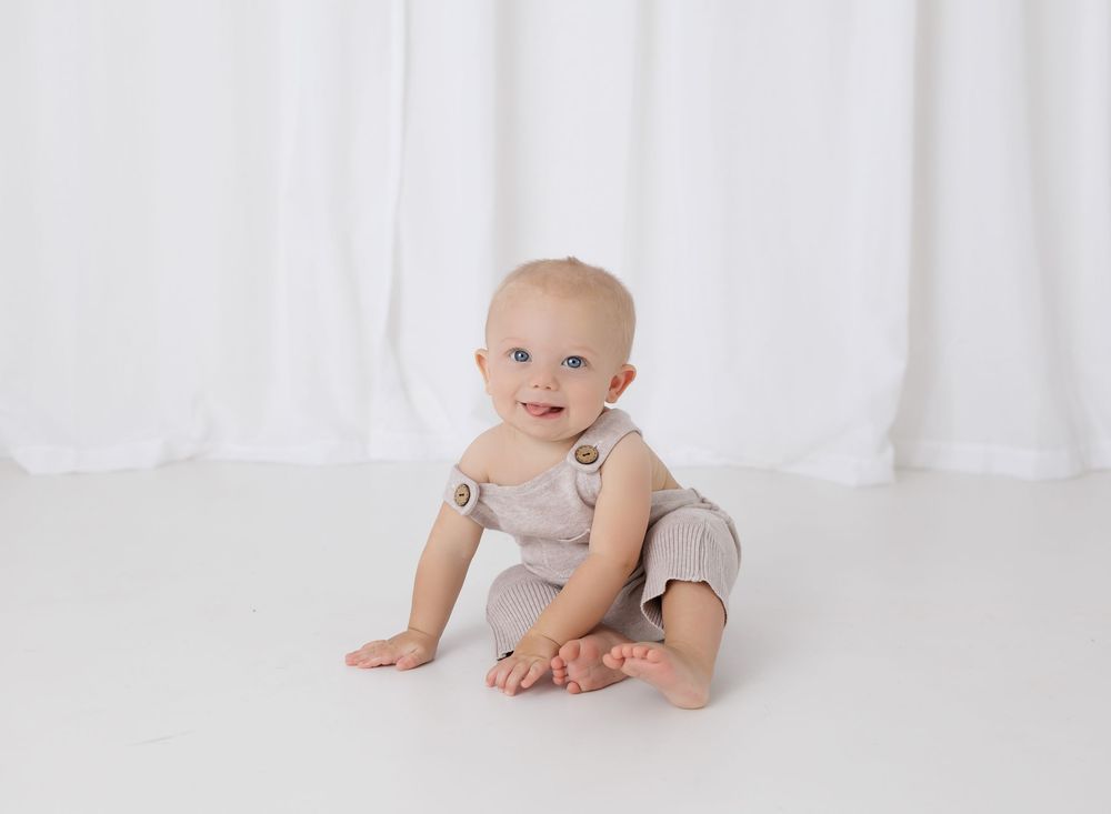 Smiling baby in a beige romper sitting on a white floor against a white draped backdrop.