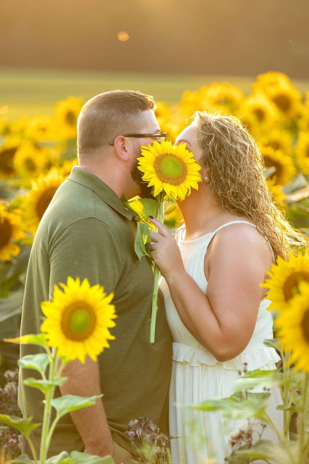 Couple sharing a romantic moment behind a sunflower in a golden sunlit field during sunset.