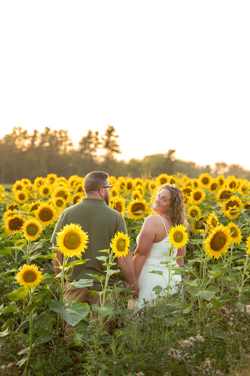 Woman in white dress looks back smiling while couple walks through sunflower field.
