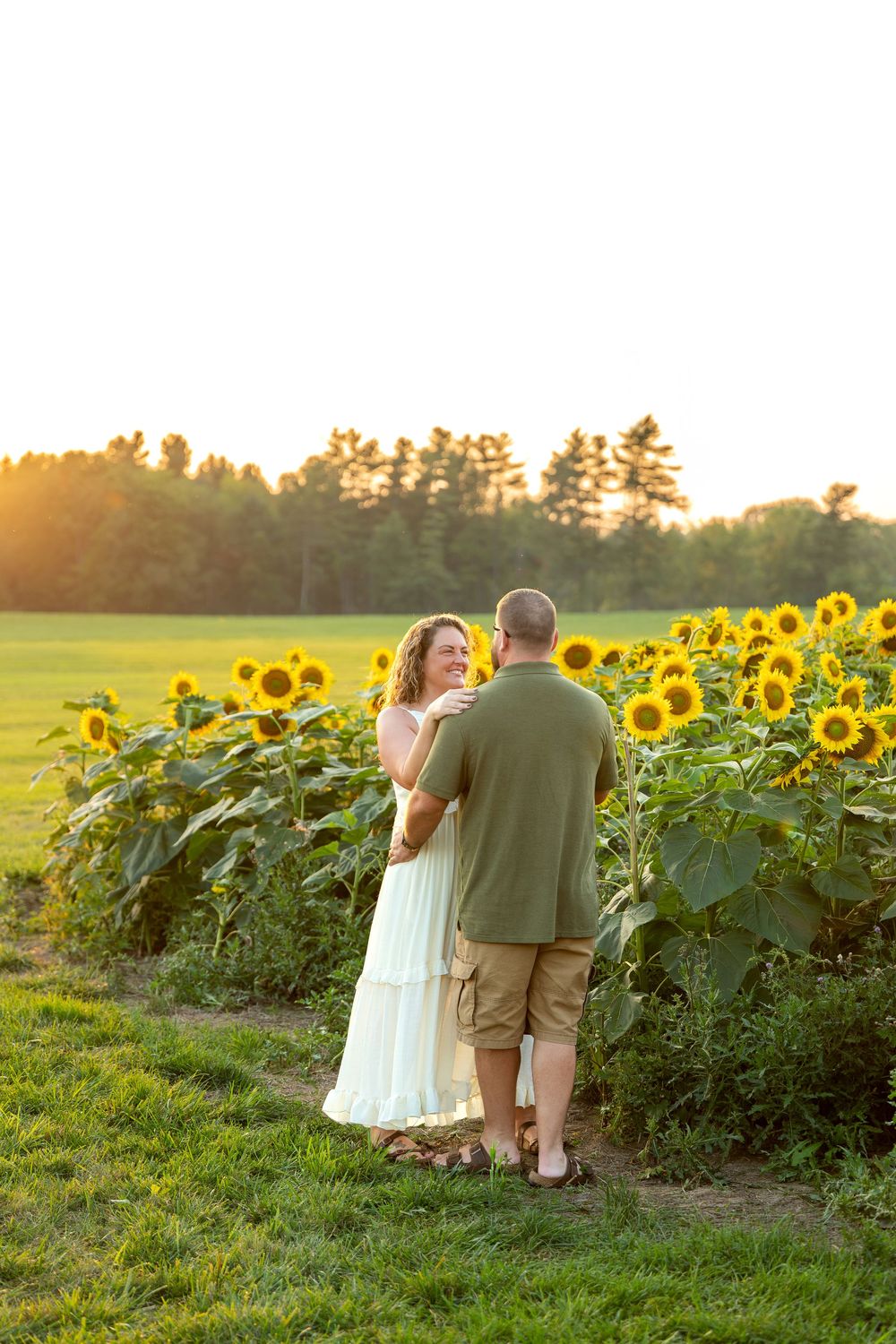 Couple dancing together in a sunflower field at golden hour, woman in white dress smiling at man in green shirt.