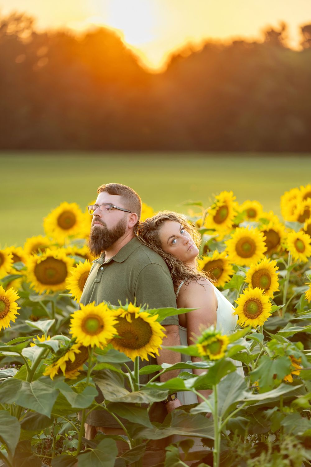 Couple standing back-to-back surrounded by blooming sunflowers at golden hour sunset.