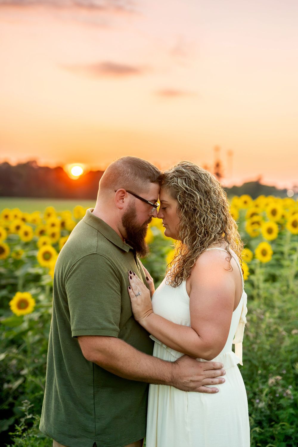 Couple embracing forehead-to-forehead in a sunflower field during a vibrant orange and pink sunset.