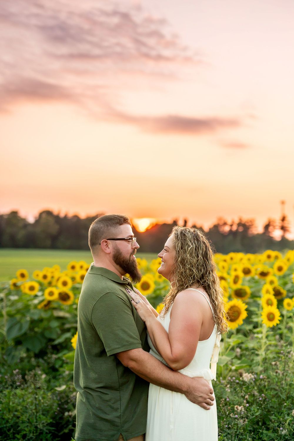Romantic couple embracing in a sunflower field during a vibrant pink and orange sunset.