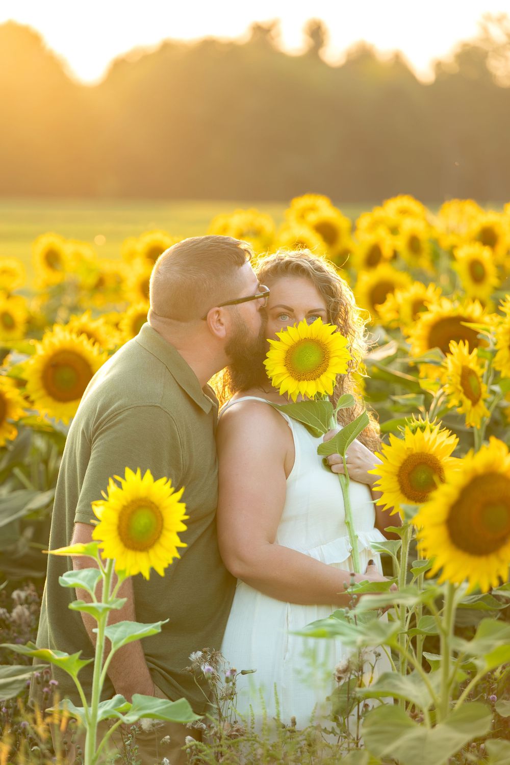 Couple embracing in a sunflower field at golden hour, man kissing woman's cheek as she holds a sunflower.