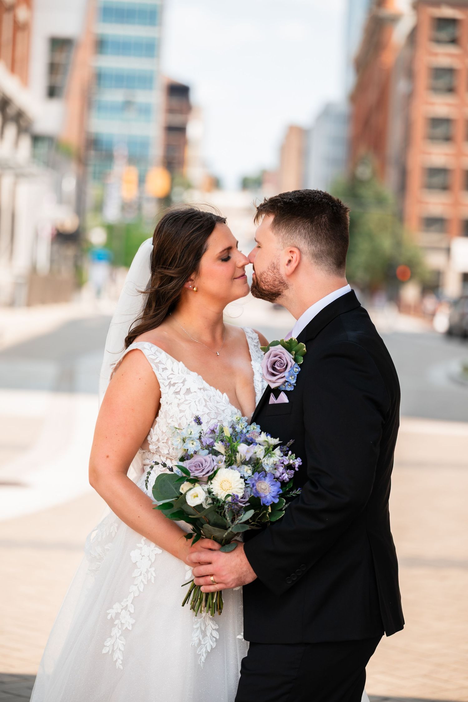 Bride and groom share a romantic nose-to-nose moment on a city street, bride holding a purple and white bouquet.