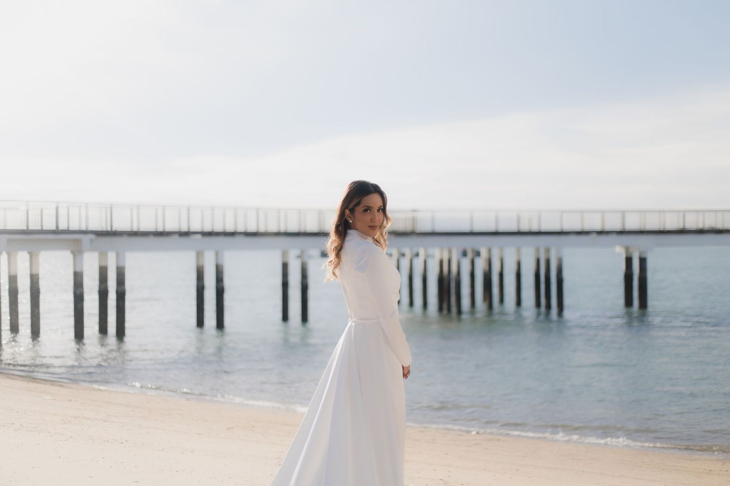 Woman in white dress posing on sandy beach near wooden pier with calm ocean water in background.