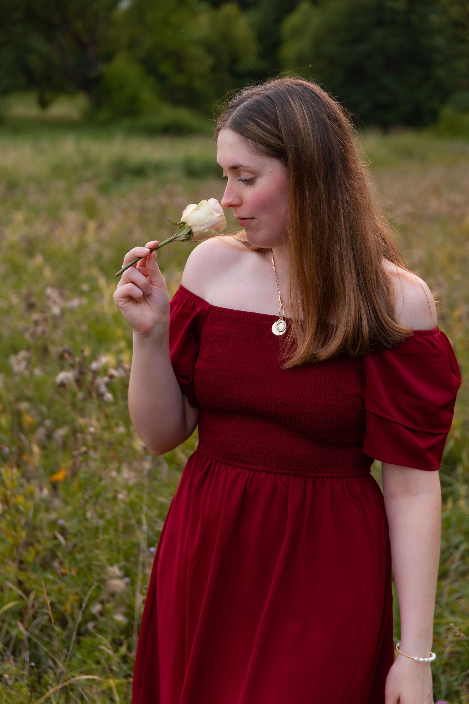 Young woman in red off-shoulder dress smelling a white flower in a green meadow field.