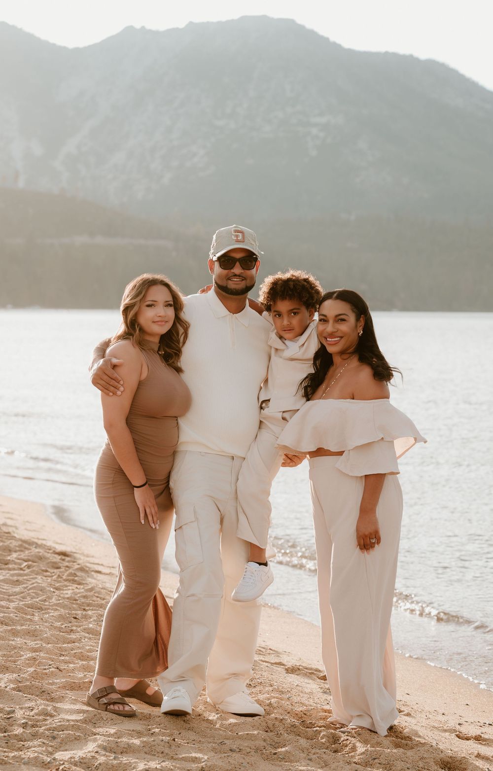 Family of four posing on a sandy beach with mountain lake backdrop, wearing coordinated neutral and white outfits.