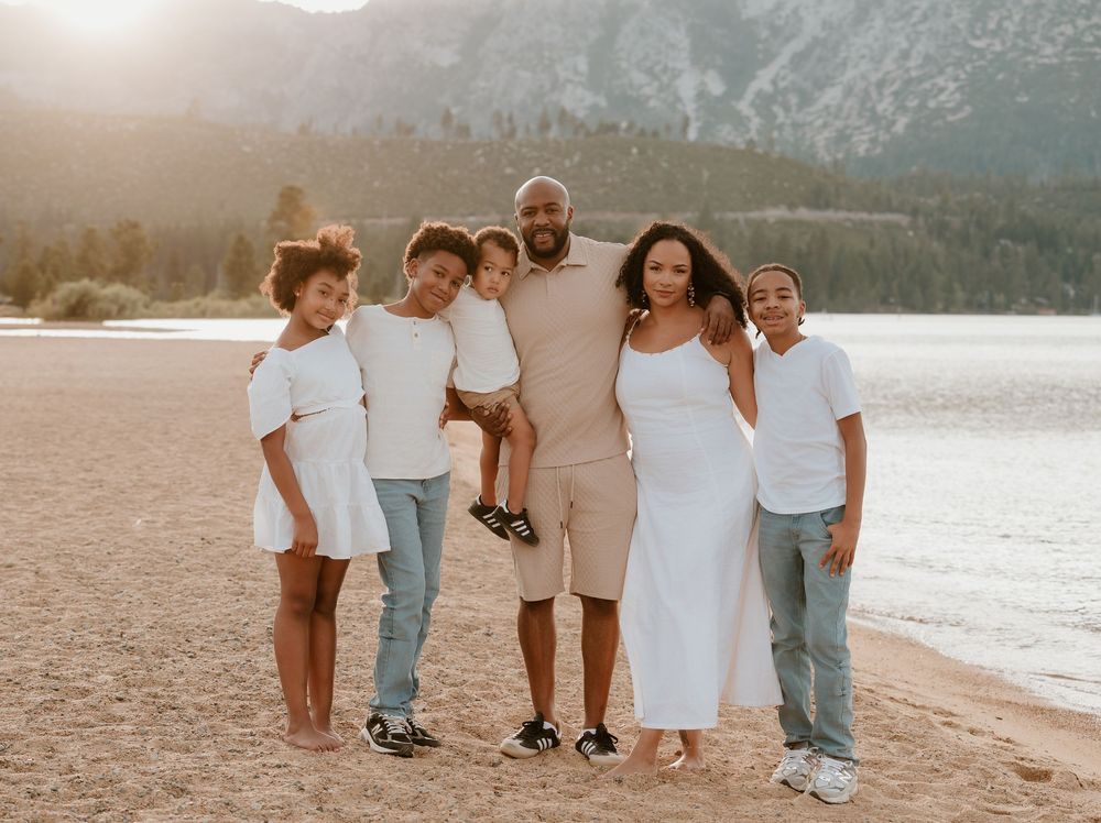 Happy family of six posing together on a sandy beach at golden hour with mountains in the background.