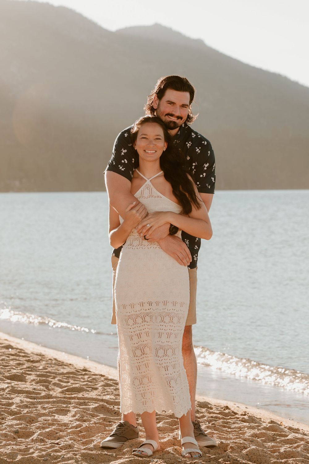 Couple embracing on a sandy beach at golden hour with mountains and calm water behind them.