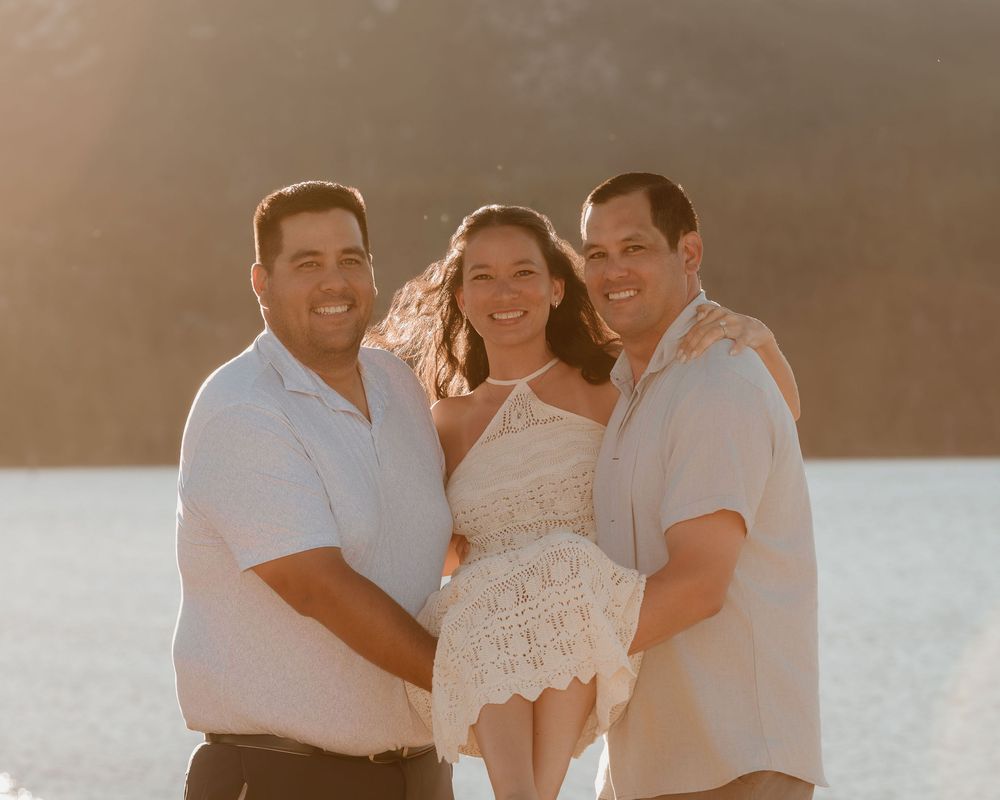 Three friends smiling together on a sunny beach with golden backlight and blue water in the background.