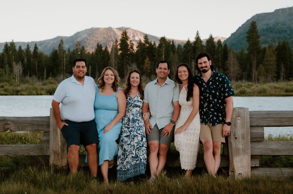 Six people smiling together outdoors near a wooden fence with mountain lake and pine trees in background.