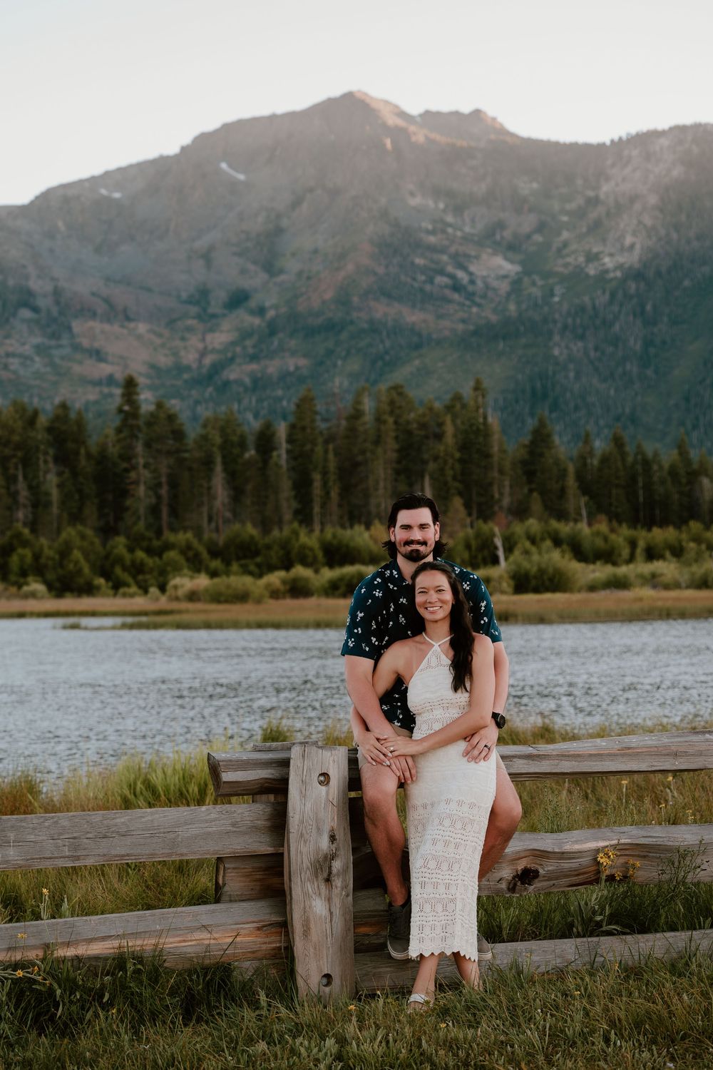 Couple poses by rustic fence with mountain lake and pine forest backdrop at golden hour.