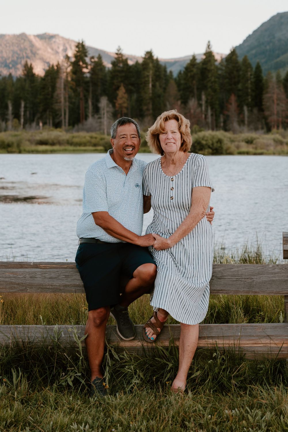 Smiling couple posing by a mountain lake with pine trees and peaks in the background on a sunny day.