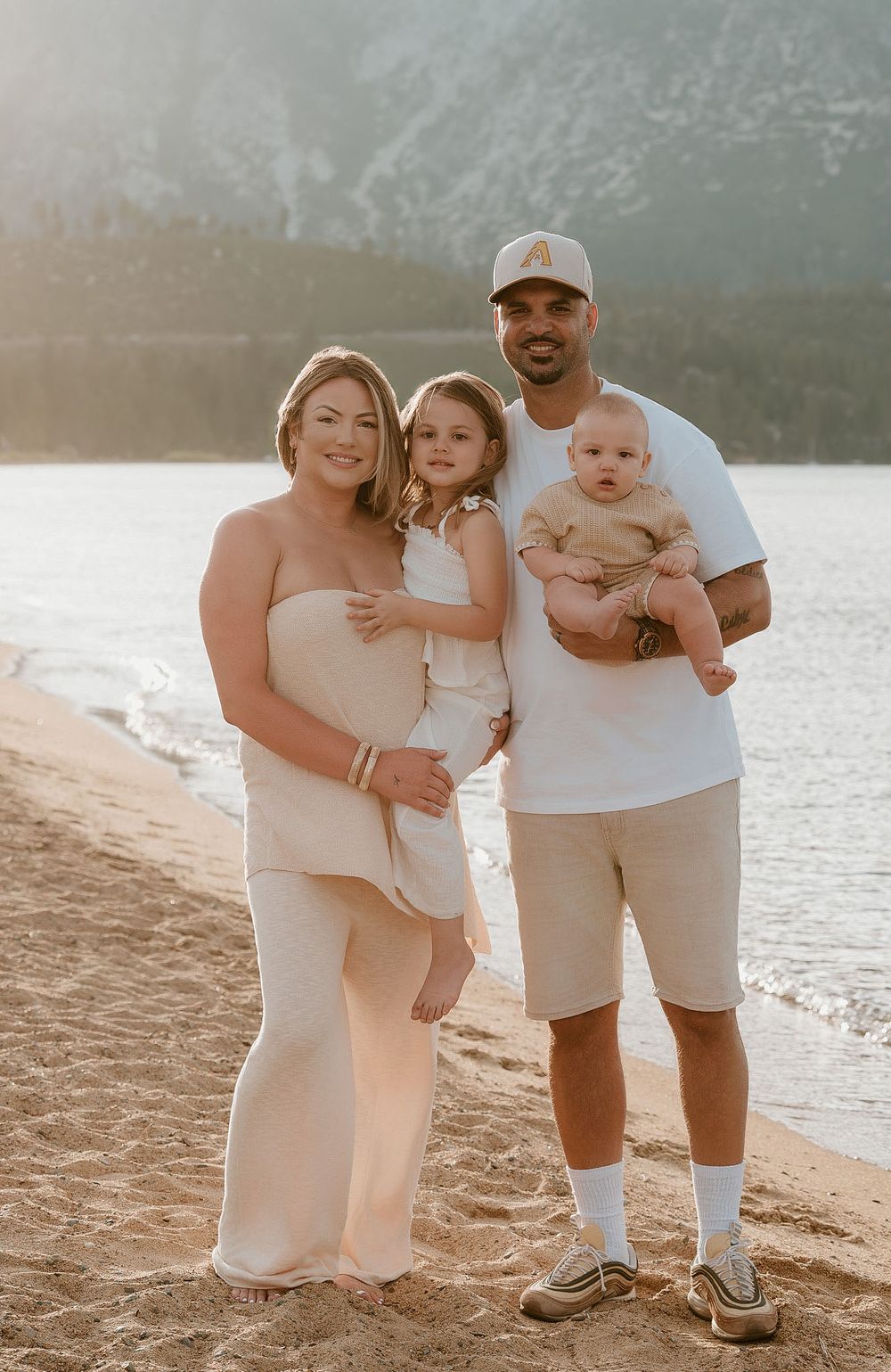 A happy family of four poses on a sandy beach at golden hour, with parents holding two young children.