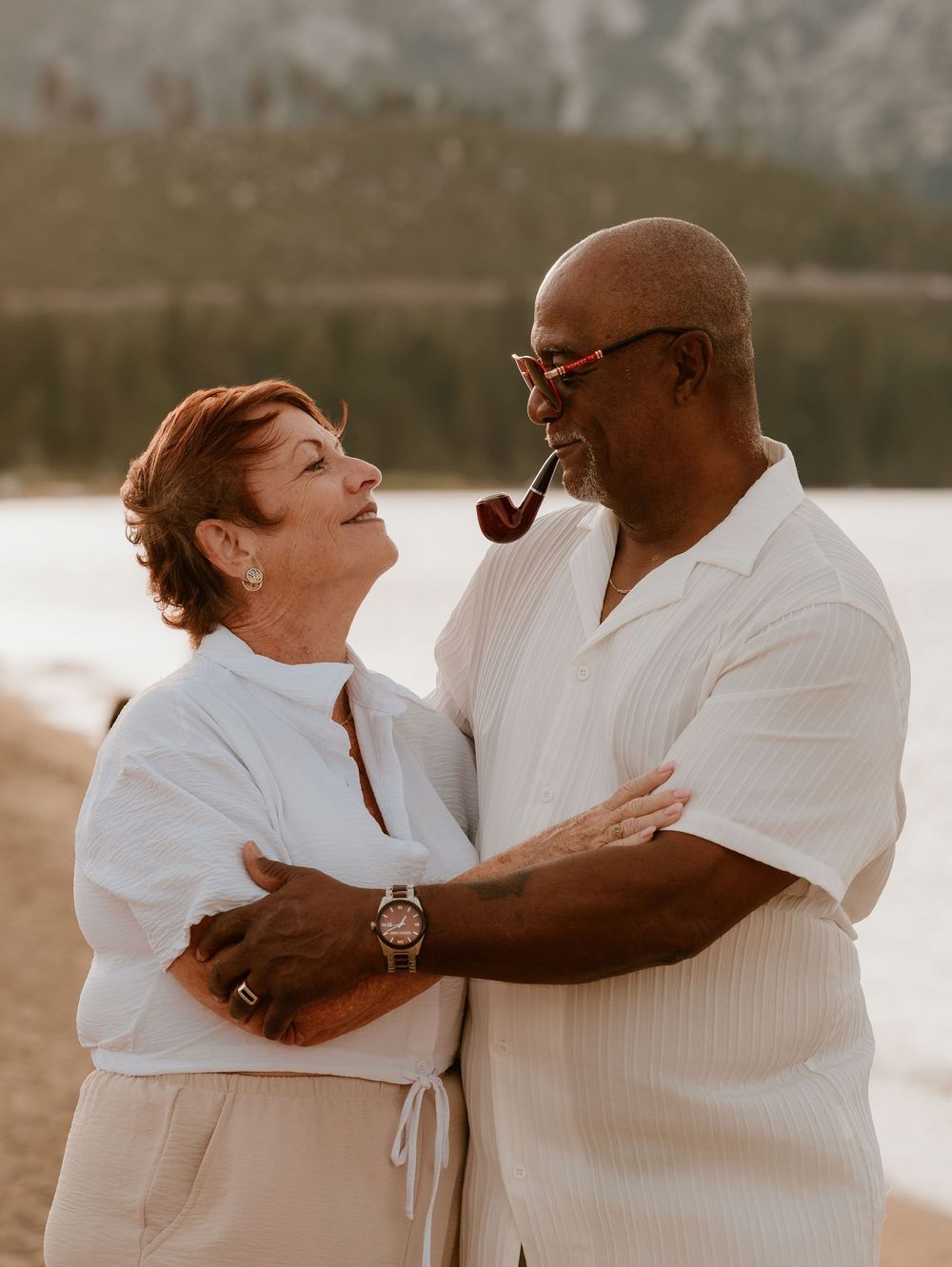 An older interracial couple embraces and smiles at each other on a lakeside beach, both wearing white outfits.