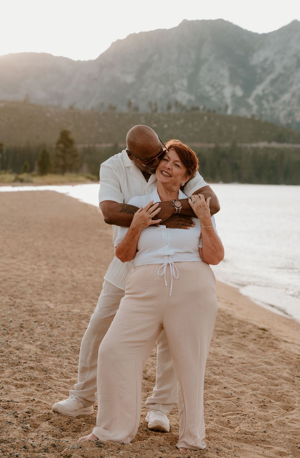 A couple embraces on a sandy beach shore with mountains and a lake in the background at golden hour.