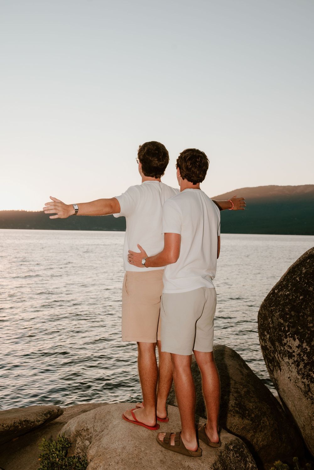 Two men standing on rocks embracing while overlooking a serene lake at sunset, one pointing toward the horizon.